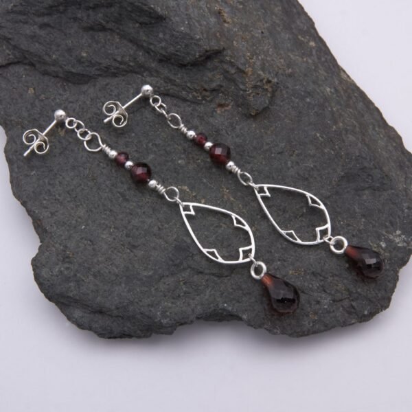 Garnet and Sterling Silver Deco Style Dangle Earrings with two smaller garnets and silver beads at the top, a Deco style silver teardrop frame below, and a teardrop shaped garnet suspended below the frame, shown laid flat on a dark grey stone
