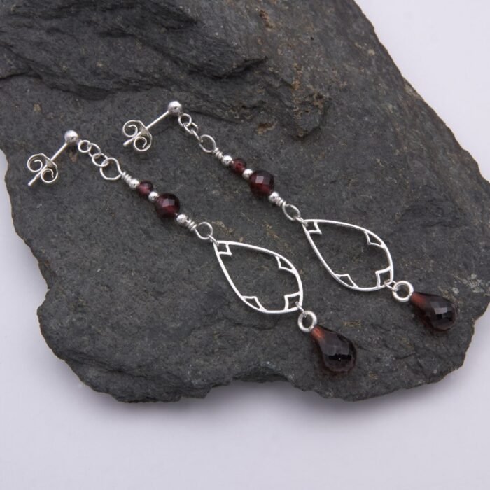 Garnet and Sterling Silver Deco Style Dangle Earrings Garnet and Sterling Silver Deco Style Dangle Earrings with two smaller garnets and silver beads at the top, a Deco style silver teardrop frame below, and a teardrop shaped garnet suspended below the frame, shown laid flat on a dark grey stone