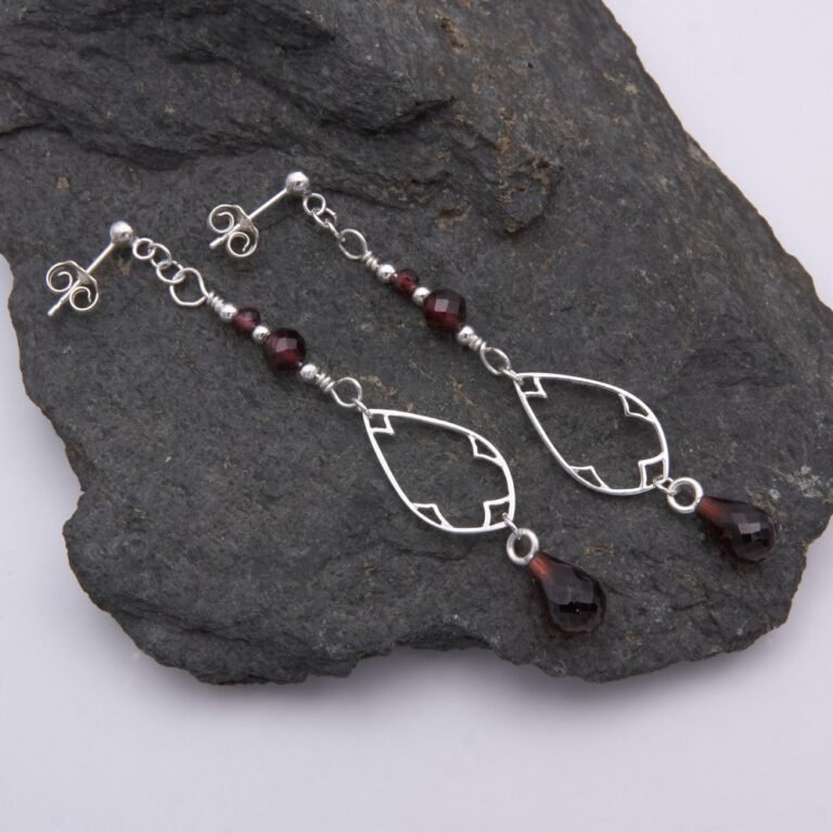 Garnet and Sterling Silver Deco Style Dangle Earrings Garnet and Sterling Silver Deco Style Dangle Earrings with two smaller garnets and silver beads at the top, a Deco style silver teardrop frame below, and a teardrop shaped garnet suspended below the frame, shown laid flat on a dark grey stone