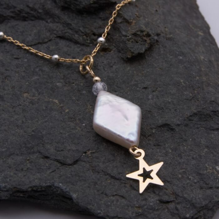 A handmade necklace featuring a diamond-shaped baroque freshwater pearl, cubic zirconia bead and gold filled star. The freshwater pearl is ivory with a pearlescent lustre and displays a variety of colours as it catches the light. Beneath the pearl is suspended a gold filled open star. A faceted round white cubic zirconia stone sits above, together with a 2mm smooth round gold filled bead. This is suspended from a gold filled satellite chain with sterling silver beads spaced evenly all around the chain. Image shows close-up of diamond and star with partial view of chain, on a dark stone background.