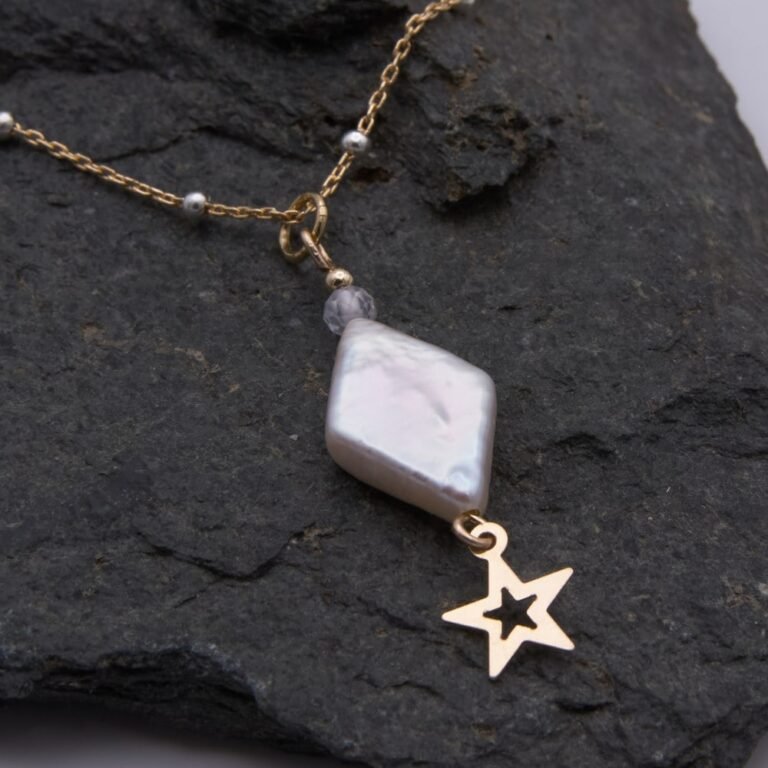 A handmade necklace featuring a diamond-shaped baroque freshwater pearl, cubic zirconia bead and gold filled star. The freshwater pearl is ivory with a pearlescent lustre and displays a variety of colours as it catches the light. Beneath the pearl is suspended a gold filled open star. A faceted round white cubic zirconia stone sits above, together with a 2mm smooth round gold filled bead. This is suspended from a gold filled satellite chain with sterling silver beads spaced evenly all around the chain. Image shows close-up of diamond and star with partial view of chain, on a dark stone background.