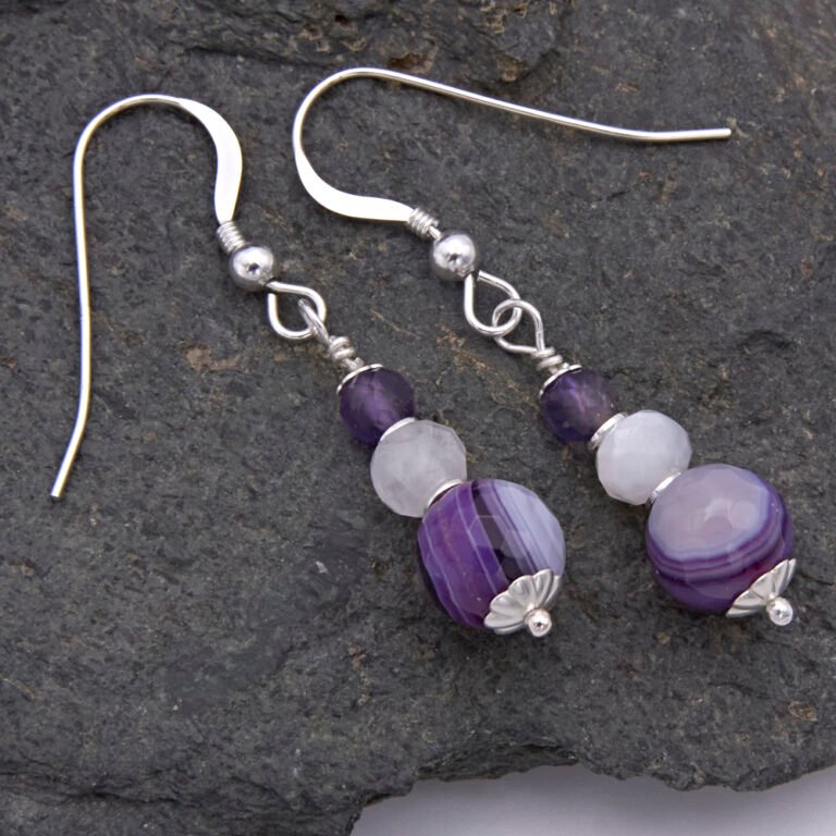 Purple Banded Agate, Rainbow Moonstone and Amethyst Sterling Silver Drop Earrings Purple Banded Agate, Rainbow Moonstone and Amethyst Stack Drop Earrings with Sterling Silver accents, suspended from Sterling Silver ear hooks, shown viewed from above on dark grey stone