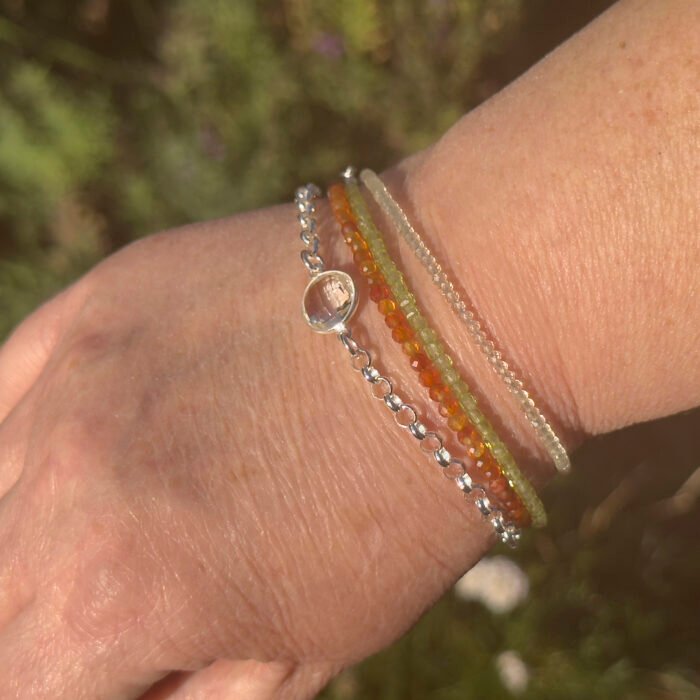Image shows Bracelet Stack including Crystal Quartz Heavyweight Belcher Chain, Lemon Quartz, Carnelian and Peridot bracelets, worn by model